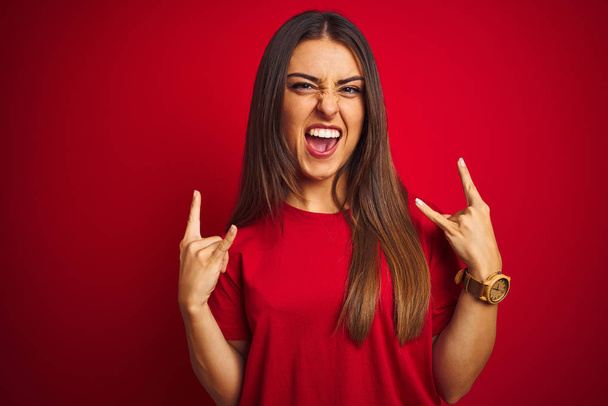 Молода красива жінка в футболці стоїть над ізольованим червоним тлом, кричить з божевільним виразом, роблячи рок-символ з руками вгору. Музична зірка. Важка концепція
. - Фото, зображення