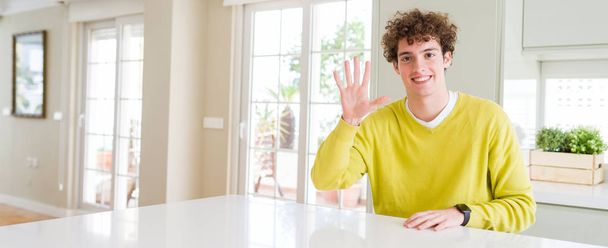 Широкоугольный снимок молодого красивого мужчины дома, показывающего и указывающего пальцами номер пять, улыбающегося уверенно и счастливо
. - Фото, изображение