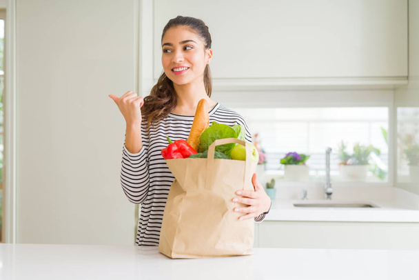 Mujer joven sosteniendo bolsa de papel llena de comestibles apuntando y mostrando con el pulgar hacia el lado con la cara feliz sonriendo
 - Foto, imagen