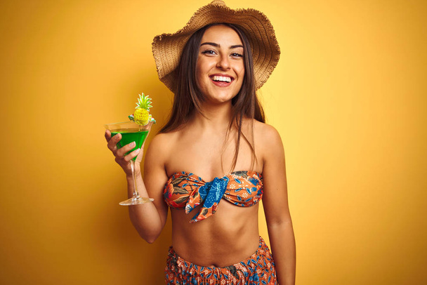Frau im Urlaub trägt Bikini und Hut und trinkt Cocktail vor isoliertem gelben Hintergrund mit einem glücklichen Gesicht stehend und lächelnd mit einem selbstbewussten Lächeln, das Zähne zeigt - Foto, Bild