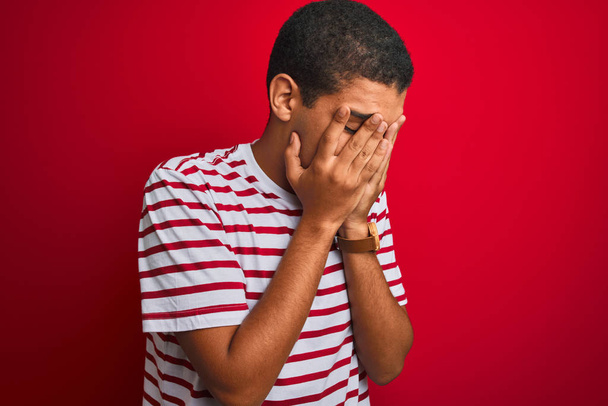 junger gutaussehender arabischer Mann in gestreiftem T-Shirt vor isoliertem roten Hintergrund mit traurigem Gesichtsausdruck, der das Gesicht mit Händen bedeckt, während er weint. Depressionskonzept. - Foto, Bild