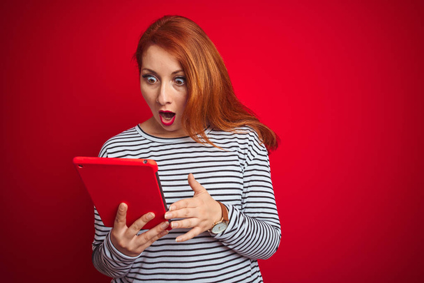 junge schöne rothaarige Frau mit Tablette über rotem isoliertem Hintergrund verängstigt im Schock mit einem Überraschungsgesicht, ängstlich und aufgeregt mit Angstausdruck - Foto, Bild
