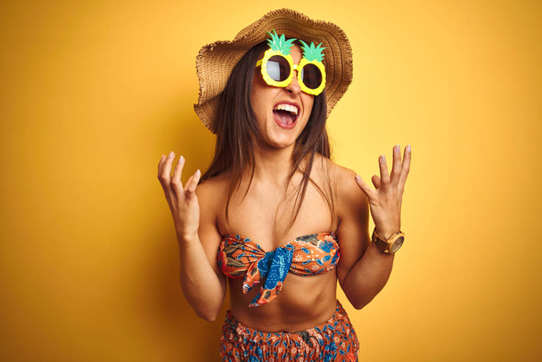 Nainen lomalla yllään bikinit ja ananas aurinkolasit yli eristetty keltainen tausta hullu ja hullu huutaa ja huutaa aggressiivinen ilme ja kädet ylhäällä. Turhautumisen käsite
. - Valokuva, kuva