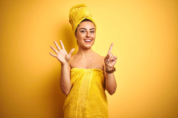 Молода красива жінка носить рушник після душу над ізольованим жовтим тлом, показуючи і вказуючи пальцями номер шість, посміхаючись впевнено і щасливо
. - Фото, зображення