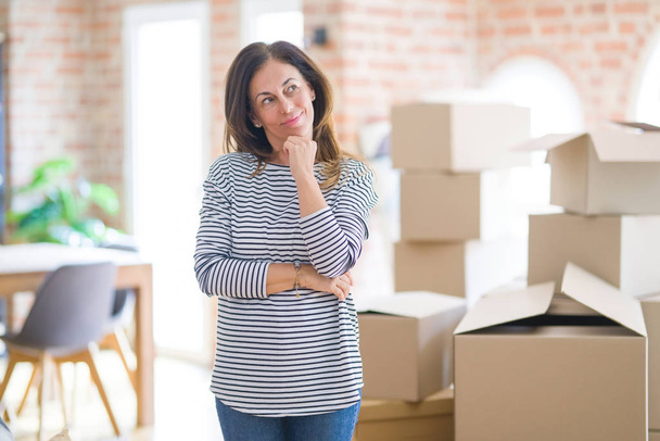Middle age woman moving to a new house arround cardboard boxes with hand on chin thinking about question, pensive expression. Smiling with thoughtful face. Doubt concept. - 写真・画像