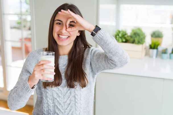 Beautiful young woman drinking a glass of fresh milk with happy face smiling doing ok sign with hand on eye looking through fingers - Фото, зображення