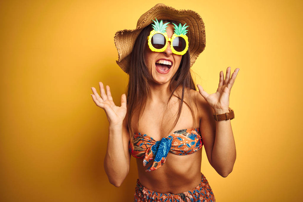 Nainen lomalla yllään bikinit ja ananas aurinkolasit yli eristetty keltainen tausta juhlii hullu ja hullu menestystä kädet kohotetaan ja suljetut silmät huutaa innoissaan. Voittajan käsite
 - Valokuva, kuva