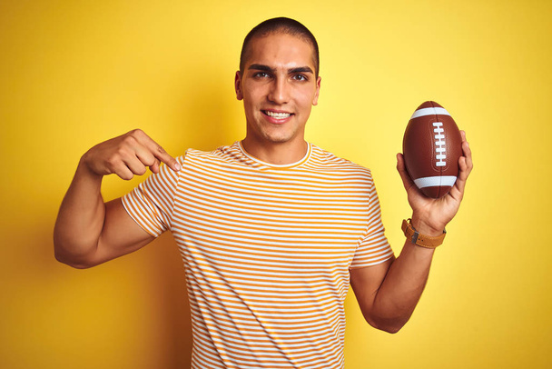 黄色の孤立した背景の上にサッカーボールを持つ若いラグビー選手の男は、自分自身に指を指して驚きの顔を持つ - 写真・画像