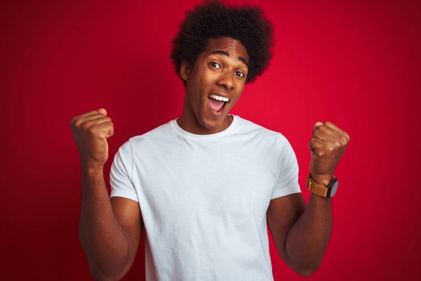 Young american man with afro hair wearing white t-shirt standing over isolated red background celebrating surprised and amazed for success with arms raised and open eyes. Winner concept. - Fotó, kép