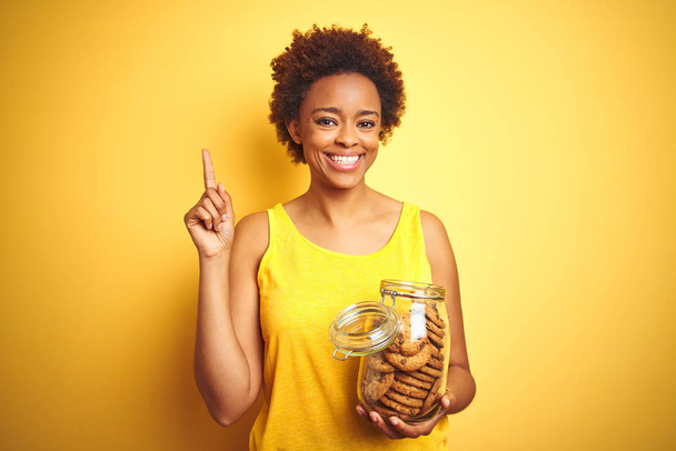 Donna afroamericana con barattolo di biscotti con gocce di cioccolato su sfondo giallo sorpresa da un'idea o domanda che punta il dito con la faccia felice, numero uno
 - Foto, immagini