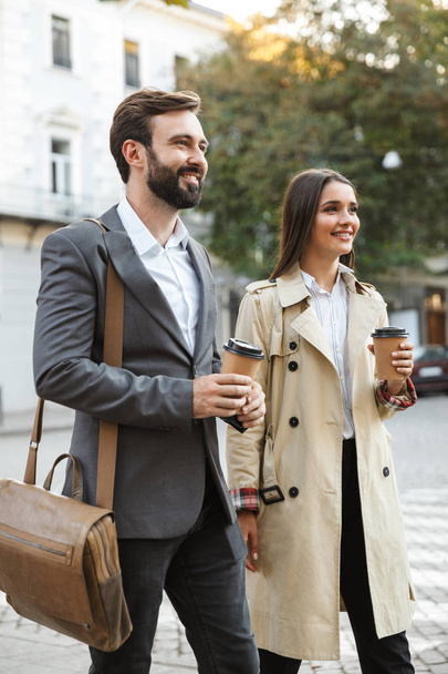 Photo of joyful office workers man and woman in formal wear drinking takeaway coffee while walking on city street - Photo, image