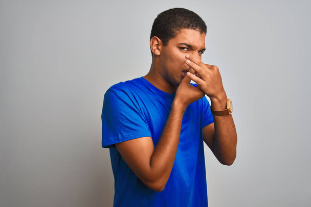 Молодий красивий арабський чоловік у синій футболці стоїть над ізольованим білим тлом, пахне чимось тьмяним і огидним, нестерпним запахом, тримаючи дихання пальцями на носі. Погані запахи концепція
. - Фото, зображення