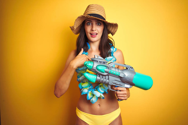 Hermosa mujer con bikini y flores hawaianas lei y pistola de agua sobre fondo aislado con la cara sorpresa señalando el dedo a sí mismo
 - Foto, imagen