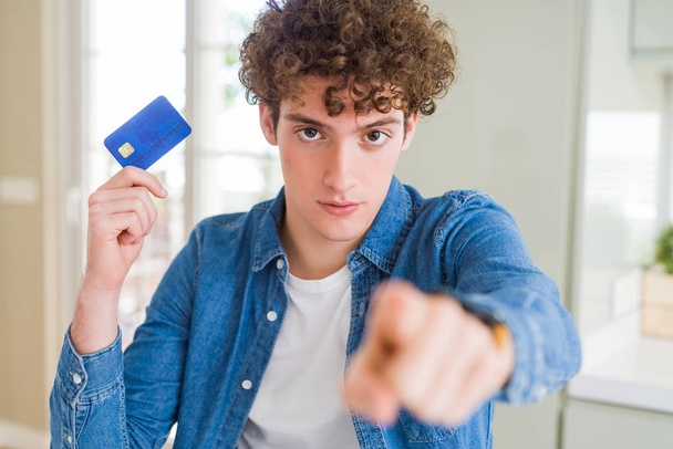 Młody człowiek trzyma kartę kredytową wskazując palcem do aparatu i do Ciebie, ręka znak, pozytywny i pewny siebie gest z przodu - Zdjęcie, obraz
