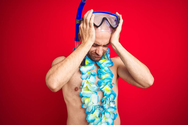 Young man wearing diving snorkel goggles and hawaiian lei flowers over isolated red background suffering from headache desperate and stressed because pain and migraine. Hands on head. - Photo, Image