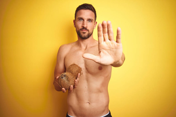Młody przystojny mężczyzna trzyma egzotyczny tropikalny kokosowy na białym tle żółty z otwartą ręką robi znak Stop z poważnym i pewnym wyrażeniem, gest obronny - Zdjęcie, obraz