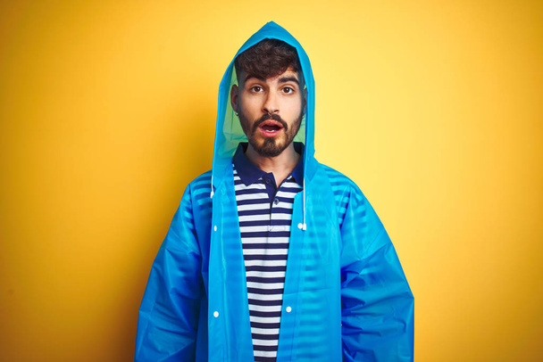 Young handsome man wearing rain coat with hood standing over isolated yellow background afraid and shocked with surprise expression, fear and excited face. - Foto, afbeelding