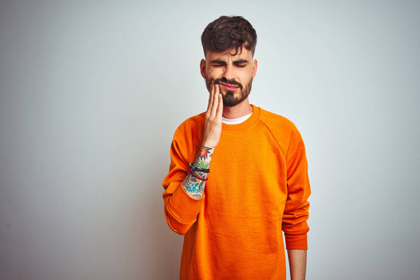 Hombre joven con tatuaje usando suéter naranja de pie sobre fondo blanco aislado tocando la boca con la mano con expresión dolorosa debido a dolor de muelas o enfermedad dental en los dientes. Concepto de dentista
. - Foto, imagen