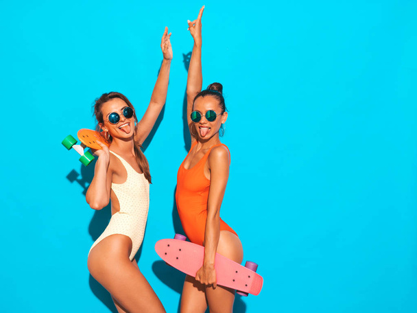 Dos hermosas mujeres hipster sonrientes sexy en trajes de baño coloridos de verano.Chicas de moda en gafas de sol. Modelos positivos que se divierten con coloridos penny skateboards.Isolated en blue.Shows lengua
 - Foto, Imagen