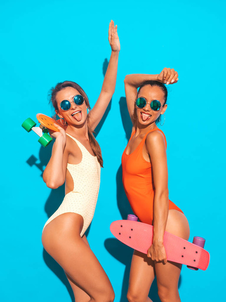 Two beautiful sexy smiling hipster women in summer colorful swimwear bathing suits.Trendy Girls in sunglasses. Positive models having fun with colorful penny skateboards.Isolated on blue.Shows tongue - Photo, Image