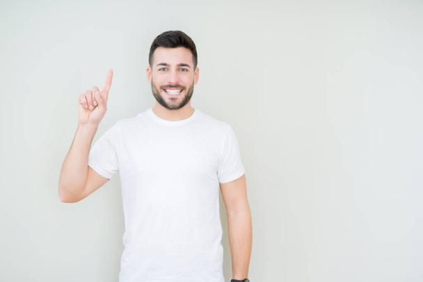 jovem bonito homem vestindo casual branco t-shirt sobre isolado fundo mostrando e apontando para cima com dedo número um enquanto sorrindo confiante e feliz
. - Foto, Imagem