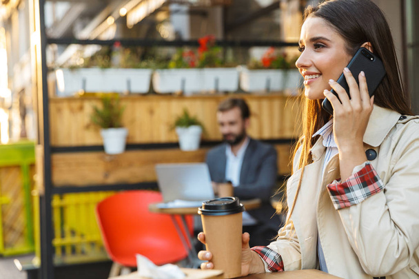 Портрет усміхненої красивої жінки, яка п'є каву і розмовляє на мобільному телефоні, сидячи в кафе
 - Фото, зображення