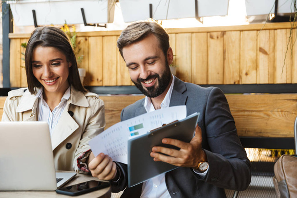 Фото усміхнених офісних працівників чоловік і жінка, які дивляться на документи і ноутбук під час використання мобільного телефону в кафе
 - Фото, зображення