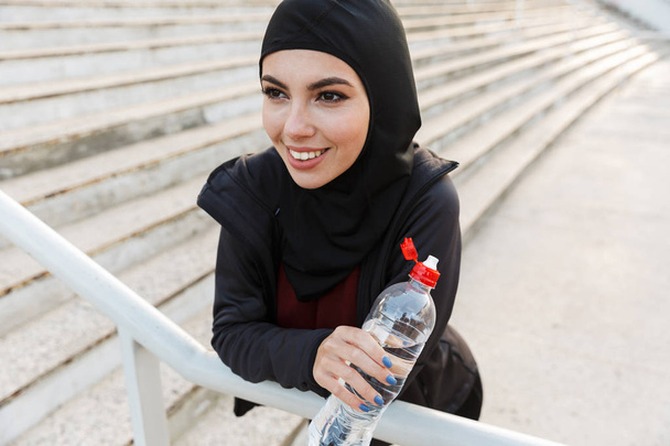 Young happy muslim sports fitness woman dressed in hijab and dark clothes outdoors at the street with steps on background drinking water holding bottle. - Photo, image