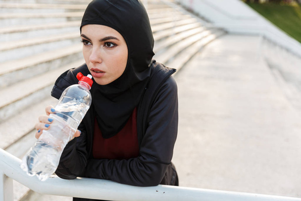 Young concentrated muslim sports fitness woman dressed in hijab and dark clothes outdoors at the street with steps on background drinking water holding bottle. - Photo, Image