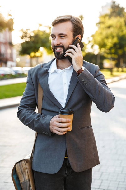 Portrait of professional adult businessman in formal suit holding takeaway coffee and talking on smartphone outdoors - 写真・画像