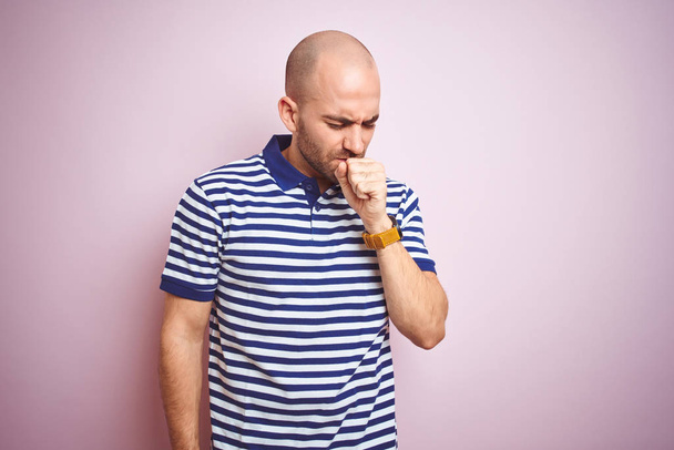 Młody łysy człowiek z brodą noszenie casual pasiasty niebieski t-shirt nad różowym białym tle uczucie złe samopoczucie i kaszel jako symptom na zimno lub zapalenie oskrzeli. Koncepcja opieki zdrowotnej. - Zdjęcie, obraz