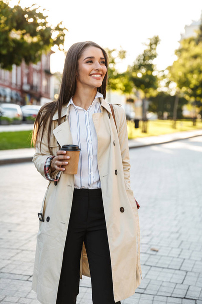 Portrait of joyful caucasian woman drinking takeaway coffee and smiling aside while walking on city street - 写真・画像