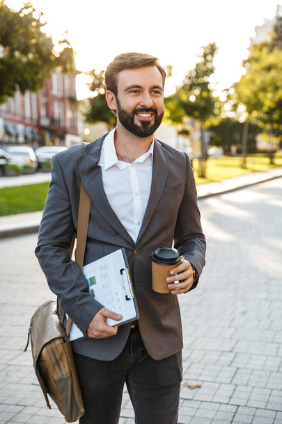 Portrait of happy adult businessman in formal suit holding takeaway coffee while walking through city street - Foto, Imagem