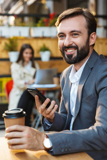 Retrato de hombre sonriente sin afeitar bebiendo café para llevar y usando el teléfono móvil mientras está sentado en la cafetería
 - Foto, imagen