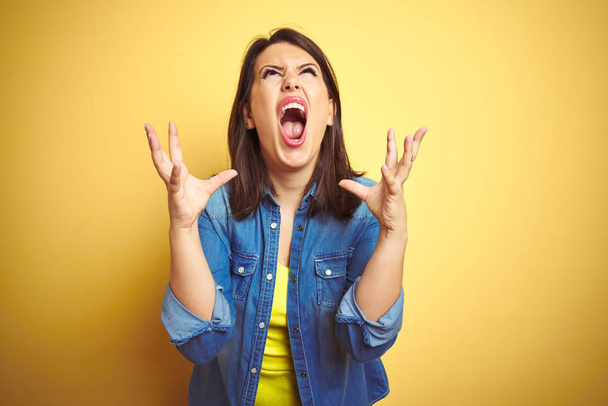 Young beautiful brunette woman wearing casual denim jacket over yellow isolated background crazy and mad shouting and yelling with aggressive expression and arms raised. Frustration concept. - Foto, afbeelding