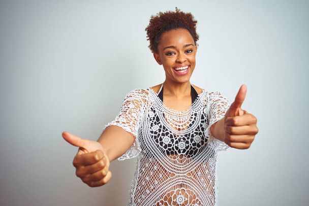 Молодая африканская американка с афроволосами в бикини на белом изолированном фоне, одобряющая позитивный жест рукой, улыбается и радуется успеху. Жест победителя
. - Фото, изображение