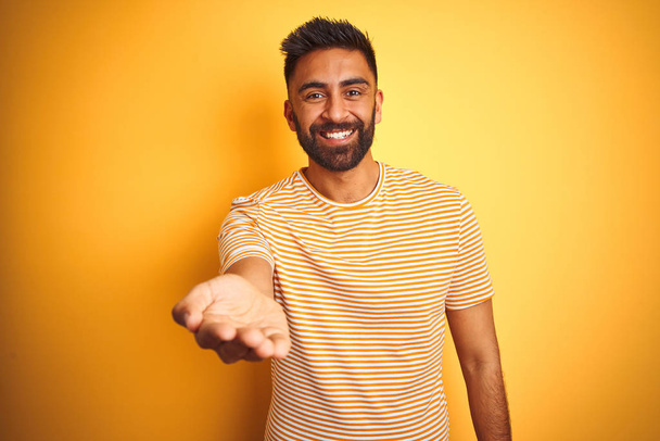 Young indian man wearing t-shirt standing over isolated yellow background smiling cheerful offering palm hand giving assistance and acceptance. - Zdjęcie, obraz