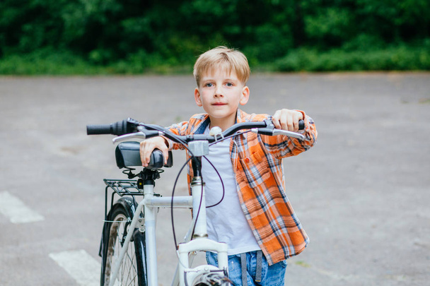 Turuncu gömlekli çocuk açık yaz parkında büyük bir bisiklet taşır. - Fotoğraf, Görsel