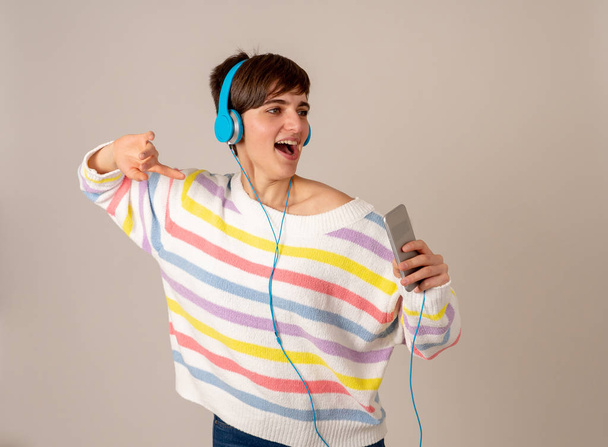 Portrait of gorgeous and happy brunette woman in colorful striped jumper listening music in headphones singing and dancing on neutral background. In positive emotions, youth and happiness concept. - Photo, Image