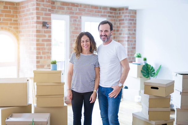 Middle age senior couple moving to a new home with boxes around with a happy and cool smile on face. Lucky person. - Photo, Image
