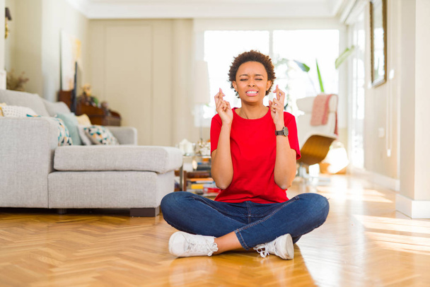 Jovem mulher americana africana bonita sentada no chão em casa sorrindo cruzando os dedos com esperança e os olhos fechados. Sorte e conceito supersticioso
. - Foto, Imagem