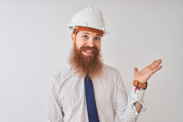 Young redhead irish architect man wearing security helmet over isolated white background smiling cheerful presenting and pointing with palm of hand looking at the camera. - Fotó, kép