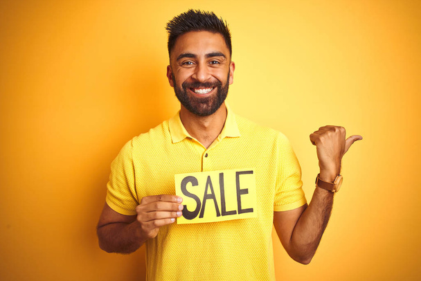 Joven árabe indio hispano sosteniendo pancarta de venta sobre aislado fondo amarillo señalando y mostrando con el pulgar hacia el lado con la cara feliz sonriendo
 - Foto, imagen
