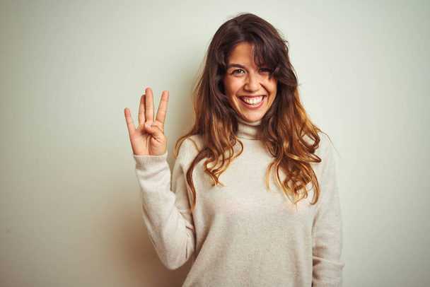 Молода красива жінка в зимовому светрі стоїть над білим ізольованим фоном, показуючи і вказуючи пальцями номер чотири, посміхаючись впевнено і щасливо
. - Фото, зображення
