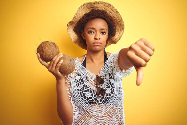 Mujer afroamericana joven con cabello afro sosteniendo coco sobre fondo aislado amarillo con cara enojada, signo negativo que muestra aversión con los pulgares hacia abajo, concepto de rechazo
 - Foto, imagen