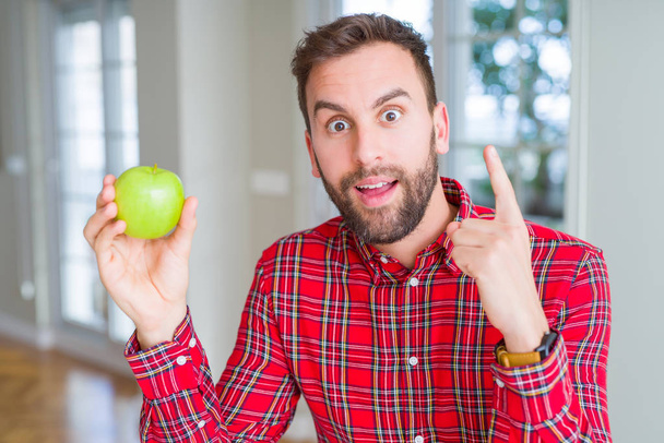Hombre guapo comiendo manzana verde fresca y saludable sorprendido con una idea o pregunta apuntando con el dedo con la cara feliz, número uno
 - Foto, Imagen