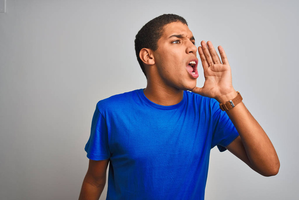 Молодий красивий арабський чоловік у синій футболці стоїть над ізольованим білим тлом і кричить голосно на бік з рукою на рот. Концепція комунікації
. - Фото, зображення