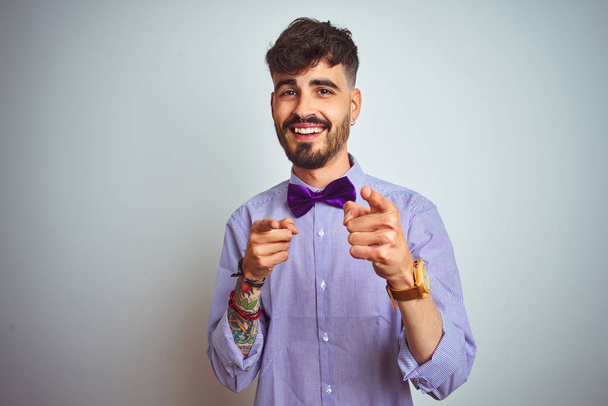 Jonge man met Tattoo dragen paarse shirt en Bow tie over geïsoleerde witte achtergrond wijzende vingers aan de camera met gelukkig en grappig gezicht. Goede energie en Vibes. - Foto, afbeelding