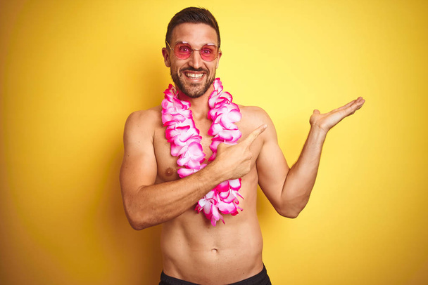 Fiatal csinos félmeztelen ember visel napszemüveget és rózsaszín Hawaii lei felett sárga háttér csodálkoztak, és mosolyogva, hogy a kamera, miközben bemutatja a kezét, és mutatva ujjal. - Fotó, kép