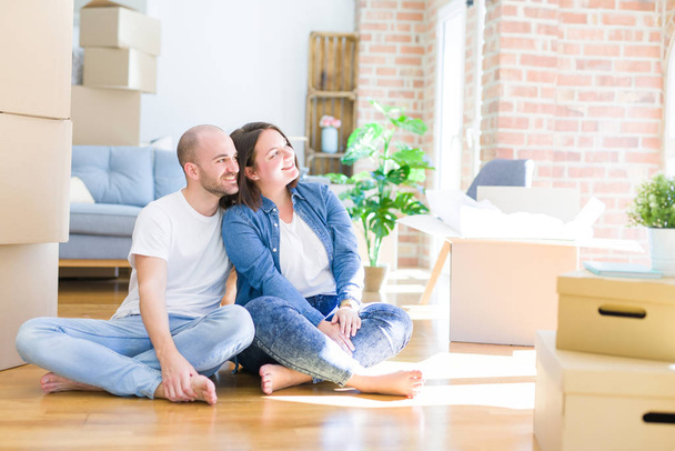 Νεαρό ζευγάρι που κάθεται στο πάτωμα πάνω από χαρτοκιβώτια που κινούνται σε ένα νέο σπίτι κοιτάζοντας μακριά με χαμόγελο στο πρόσωπο, φυσική έκφραση. Γελώντας με αυτοπεποίθηση. - Φωτογραφία, εικόνα